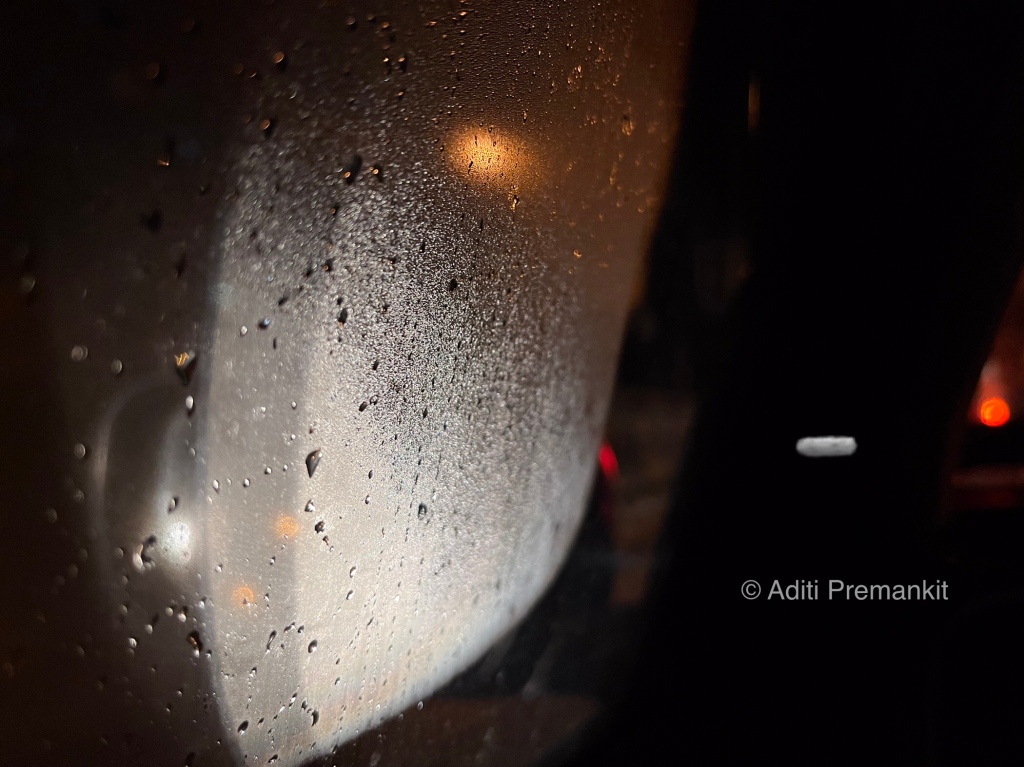 Raindrops shining on a car window while reflecting light from outside at night. 